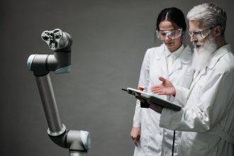 Pesquisadores usando jalecos e óculos de proteção analisam um braço robótico em um laboratório.